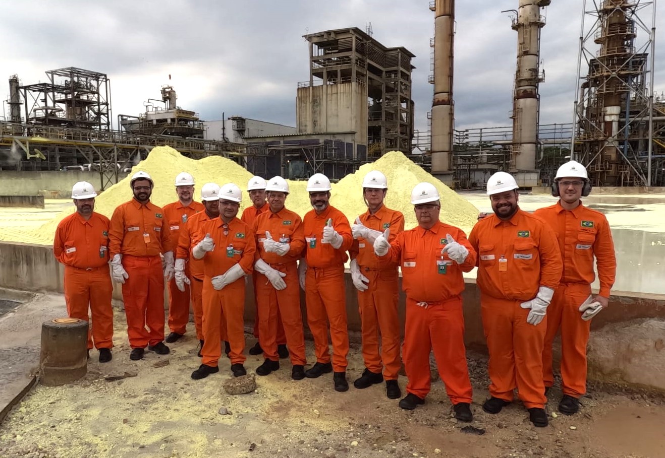Equipes de trabalhadores vestidos com macacões laranja, diante de um pátio com montes de enxofre sólido, com instalações industriais ao fundo e céu carregado de nuvens cinzas